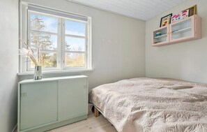 Ein Schlafzimmer mit Heizkörper, Fenster mit Blick und einem Bett mit floralem Überwurf, sowie einem rosa Schrank mit Gegenständen darauf.