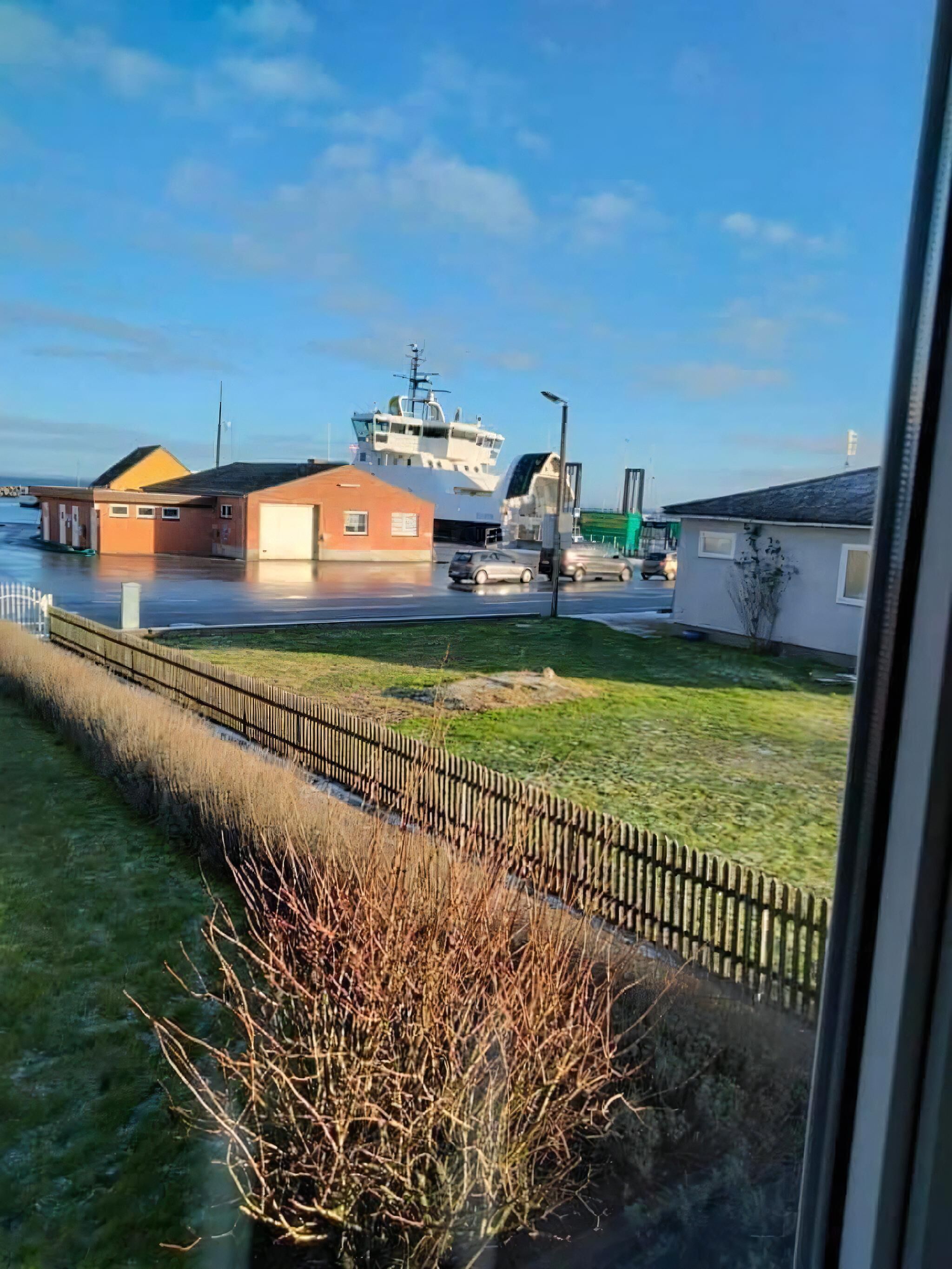 Blick auf das Wasser mit einem großen Schiff im Hafen, Gebäuden und einer grünen Fläche.