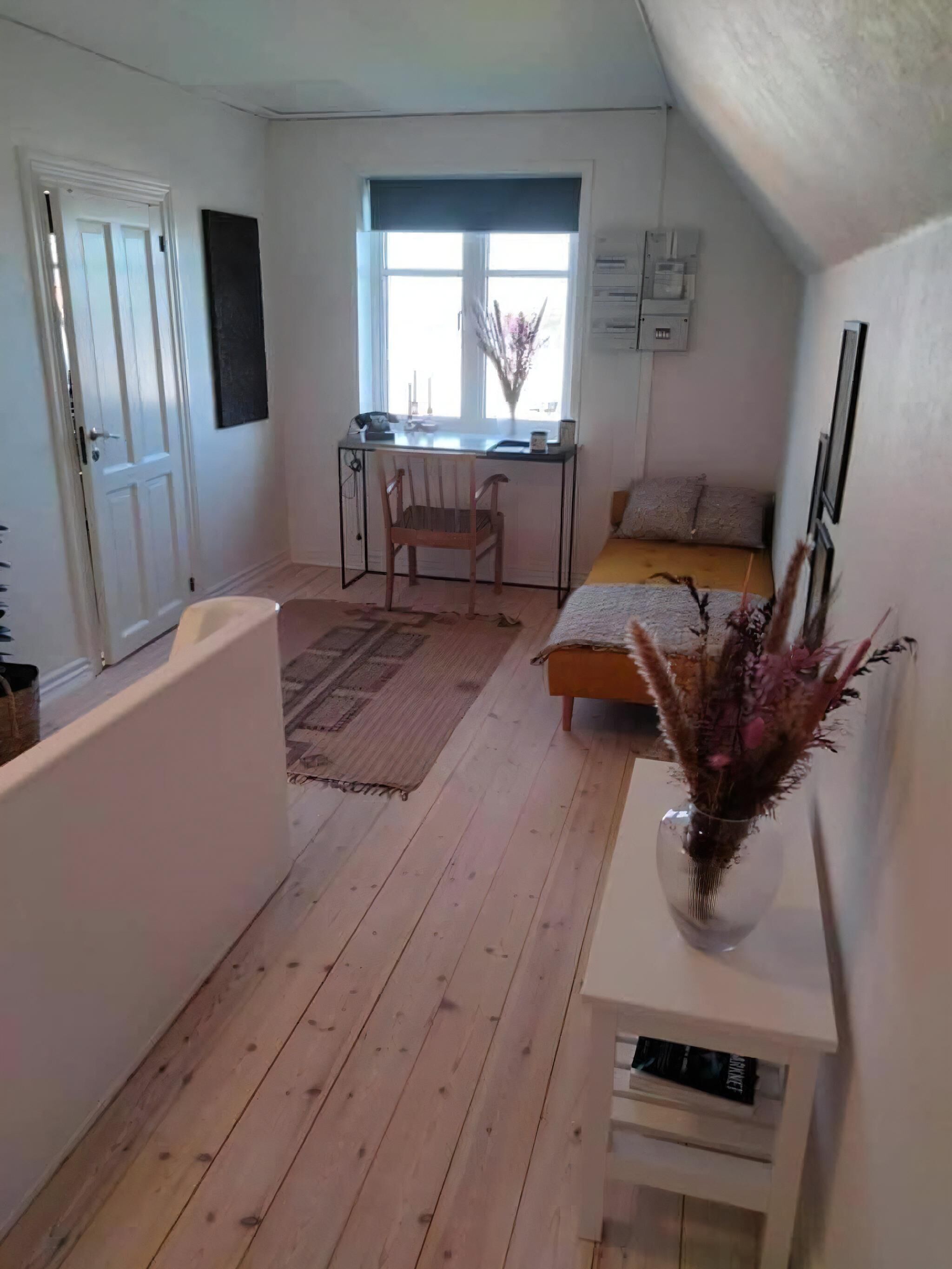 Et lille værelse med seng, skrivebord og stol. Der er et vindue med gardiner, en dør og et lille bord med en vase.