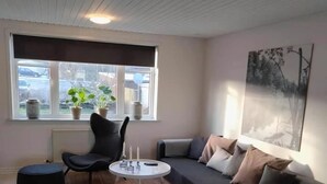 En moderne stue med en hjørnesofa, et rundt sofabord, en sort lænestol, en lille skammel og et vindue med plantekrukker.