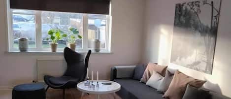 Una sala moderna con un sofá modular, una mesa de centro redonda, un sillón negro, un pequeño banquito y una ventana con plantas en macetas.