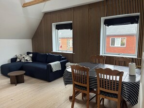 Et moderne loftværelse med en mørkeblå sofa, et spisebord med stole og store vinduer med sorte gardiner.