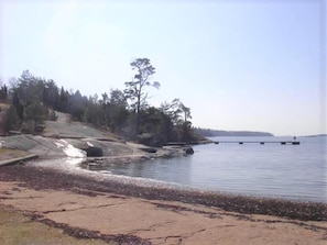 A beach with a rocky shoreline, a pier extending into calm water, and a forested area in the background. - Panoramic sea-view from the house (Bräkne-Hoby)