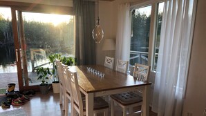 Uma sala de jantar com mesa de madeira, cadeiras brancas, luminária pendente e vista para uma floresta através de amplas janelas.