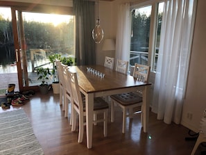 Un espace repas avec une table en bois, des chaises blanches, un luminaire suspendu et une vue sur une forêt, visible à travers de grandes fenêtres.