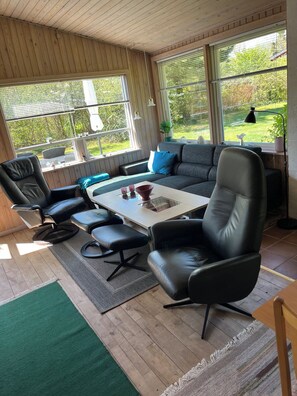 Ein modernes Wohnzimmer mit Holzbalkendecke, großen Fenstern, einem grauen Sofa, schwarzen Ledersesseln und einem kleinen Tisch mit Dekoration.