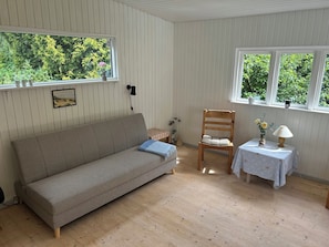 A room with a sofa, a wooden chair, a small table with a lamp, and a window with a view of greenery. - Holiday home in peaceful natural surroundings (Nørre Snede)