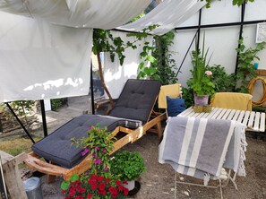 Ett takförsett utomhusområde med sittplatser, bestående av två solstolar, ett litet bord med en randig duk och krukväxter.