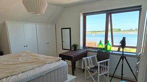Een slaapkamer met een bed, een bureau, een stoel, een statief en een raam met uitzicht op zee.