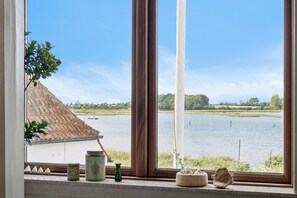 Ein Fenster mit Blick auf eine ruhige Seenlandschaft, ein Gebäude mit rotem Ziegeldach und eine Pflanze auf der Fensterbank.