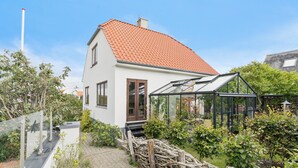 Een twee verdiepingen tellend huis met een rode dakbedekking, een glazen conservatorium en een tuin met diverse planten.