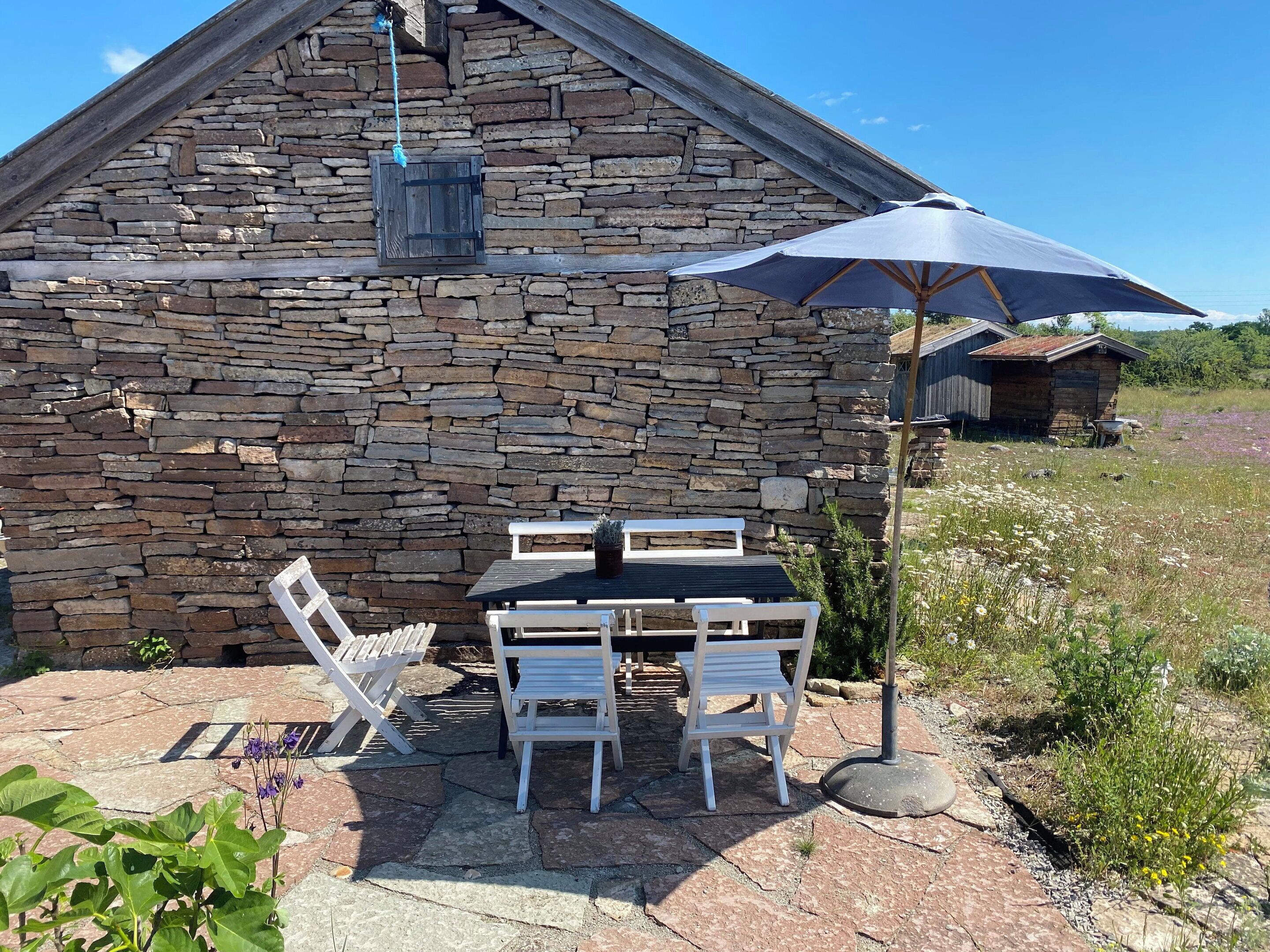 Un bâtiment en pierre avec une table et des chaises en bois, un luminaire suspendu et un parasol de patio.