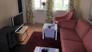 En stue med fjernsyn, en rød sofa, et hvidt bord med lampe og et vindue med gardiner.