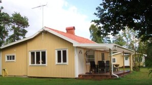 Uma casa térrea com telhado vermelho, varanda com cadeiras e um pequeno jardim.