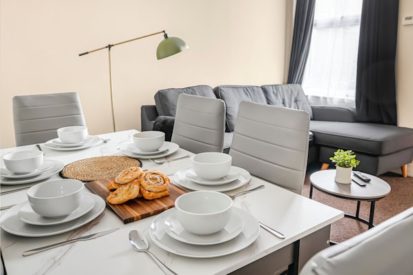 Uma área de refeições com uma mesa posta para quatro pessoas, incluindo pratos, tigelas e talheres. Há uma pequena mesa redonda com uma planta e um controle remoto. Um sofá e uma luminária são visíveis ao fundo.
