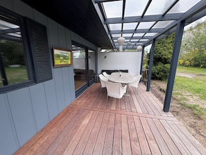 A covered patio with a wooden deck, a round table with chairs, and a built-in bench. - Marvelous views hygge in a cosy summer cottage (Asnæs)
