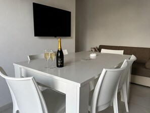A dining table with white chairs, a bottle of champagne, and two glasses. A flat-screen TV is mounted on the wall above the table.