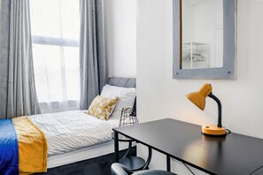 Ein Schlafzimmer mit Bett, Schreibtisch, Stuhl, Schreibtischlampe, einem Fenster mit Vorhängen und einem Spiegel an der Wand.