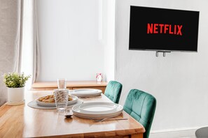 Ein Essbereich mit einem Holztisch, zwei Stühlen, Tellern, Gläsern und einer Topfpflanze. Ein an der Wand befestigter Fernseher zeigt 'NETFLIX'.