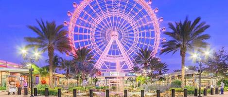Une grande roue avec une plateforme centrale offrant une vue panoramique, entourée de palmiers et bordée d’une promenade pavée.