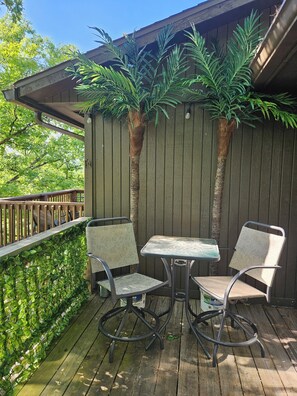 Um deck de madeira com duas cadeiras de metal e uma mesa, cercado por uma cerca de madeira e vegetação verde.