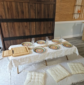 Een eettafel gedekt met borden, glazen en een houten dienblad, omgeven door een witte tafelkleed en comfortabele zitplaatsen.