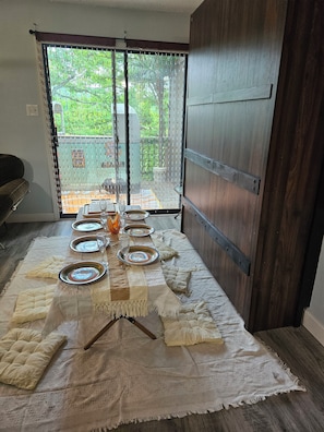 Uma área de refeições com uma mesa posta para seis pessoas, uma porta de madeira e vista para o exterior com vegetação.