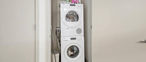 A compact laundry room with a stacked washer and dryer, a mop, and a trash bin.