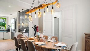 A dining area with a long table, chairs, and hanging light fixtures. - Gîtes de France® - Le Secret Des Cordelières (NEUFCHATEL EN BRAY)