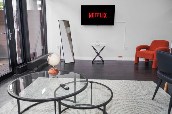En moderne stue med et glasbord, en globus, en fjernbetjening, en rød lænestol, en sort stol, et fjernsyn der viser 'NETFLIX' og et lille sofabord.
