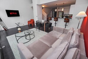 Una sala moderna con sofá, una mesa de centro, un televisor mostrando 'NETFLIX', una zona de comedor con sillas y una cocina al fondo.