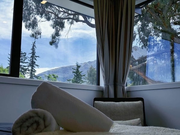 Een slaapkamer met een groot raam, een bed met kussens en uitzicht op bomen en bergen.