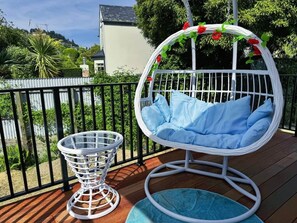 Een witte hangstoel met blauwe kussens op een houten terras, een witte rieten stoel met een blauwe kussen, en een witte mand op het terras.
