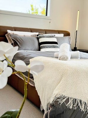 Een moderne slaapkamer met een bed, kussens, een bedlampje en een raam met uitzicht op bomen.
