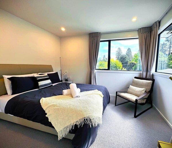 Ein modernes Schlafzimmer mit einem großen Bett, einem Sessel und einem Fenster mit Blick auf Bäume.