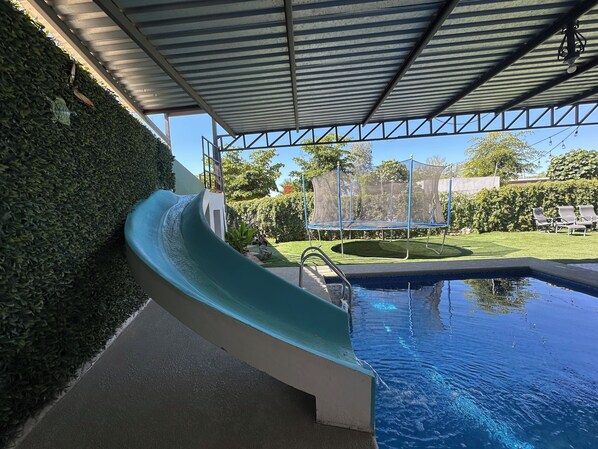 Une piscine avec un toboggan, un trampoline et un espace abrité doté d’un toit en métal.
