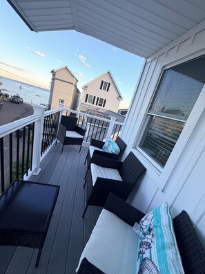 A covered outdoor deck with wicker furniture, a view of the ocean, and a house in the background.