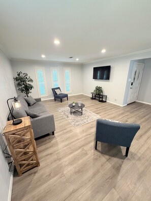 Ein geräumiges Wohnzimmer mit grauem Sofa, blauem Sessel, Couchtisch aus Holz und an der Wand montiertem Fernseher.