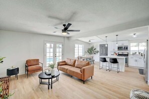 Ein modernes Wohnzimmer mit Sofa, Couchtisch und Sesseln, eine Küche mit weißen Schränken und Geräten sowie ein Deckenventilator.