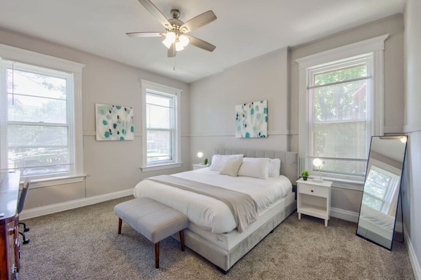 Une chambre à coucher comprenant un lit, des tables de chevet, un banc, un ventilateur de plafond et des œuvres d’art accrochées aux murs.