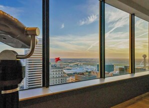 Ein modernes Interieur mit großen Fenstern mit Blick auf eine Stadtlandschaft, ein metallenes Objekt an der Fensterbank und eine Ansicht von Gebäuden sowie ein rotes Flugzeug.