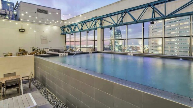 Piscina interna com vista para a paisagem urbana, deck de madeira com cadeiras e estrutura metálica treliçada.