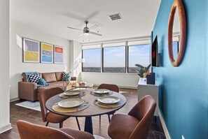 Ein modernes Wohnzimmer mit Essbereich, Deckenventilator, großen Fenstern und Wandkunst.