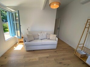 A room with a white sofa, wooden flooring, a hanging light fixture, and a window with an open door. - 250 – Cozy house with enclosed garden, close to the center, everything within walking distance (SOULAC SUR MER)