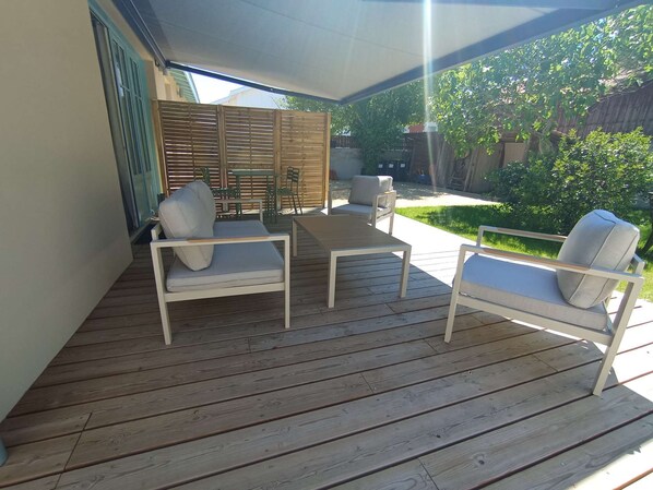 A covered outdoor patio with wooden flooring, two white cushioned chairs, a small table, and a wooden fence in the background. - 250 – Cozy house with enclosed garden, close to the center, everything within walking distance (SOULAC SUR MER)