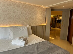 Ein Hotelzimmer mit einem Bett, Kissen, einer Decke und einem Handtuch. Eine Tür führt zu einem Badezimmer und einer Küchenzeile.