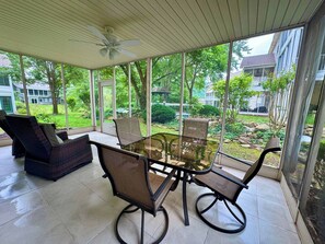 Eine überdachte Terrasse mit einem Glastisch und Stühlen, einem Deckenventilator und Blick auf eine Wohngegend mit Bäumen und Rasen.