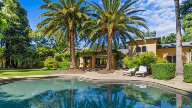 Una piscina con palmeras, un porche cubierto y un edificio de fachada color crema.