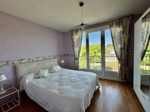 A bedroom with a bed, a nightstand, a lamp, a balcony with curtains, and a view of greenery outside. - 2-star rated house for 10 people in Mimizan (MIMIZAN)
