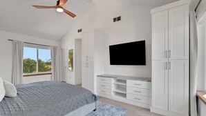 Een slaapkamer met een bed, een plafondventilator, een televisie, witte kasten en een raam met gordijnen.